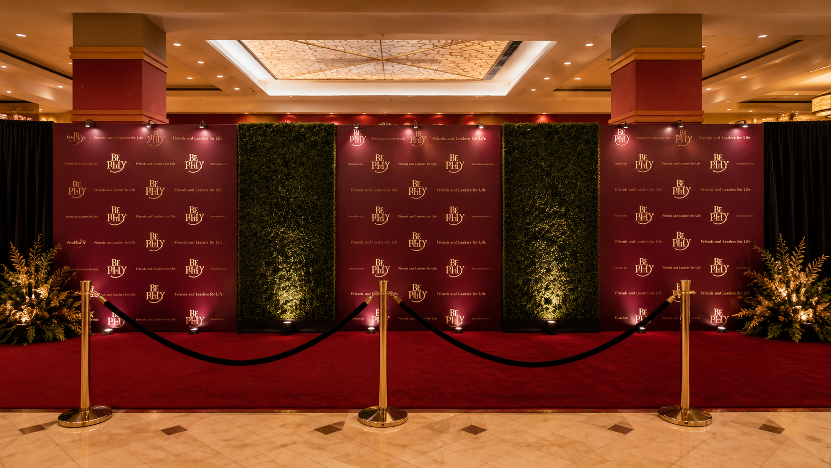Luxury custom step-and-repeat backdrop with red carpet, gold stanchions, and hedge wall accents